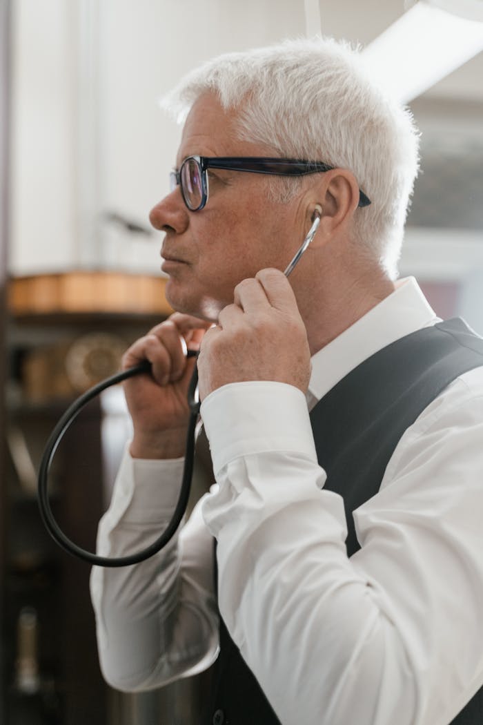 Side view of senior doctor listening with stethoscope, wearing eyeglasses and formal attire indoors.